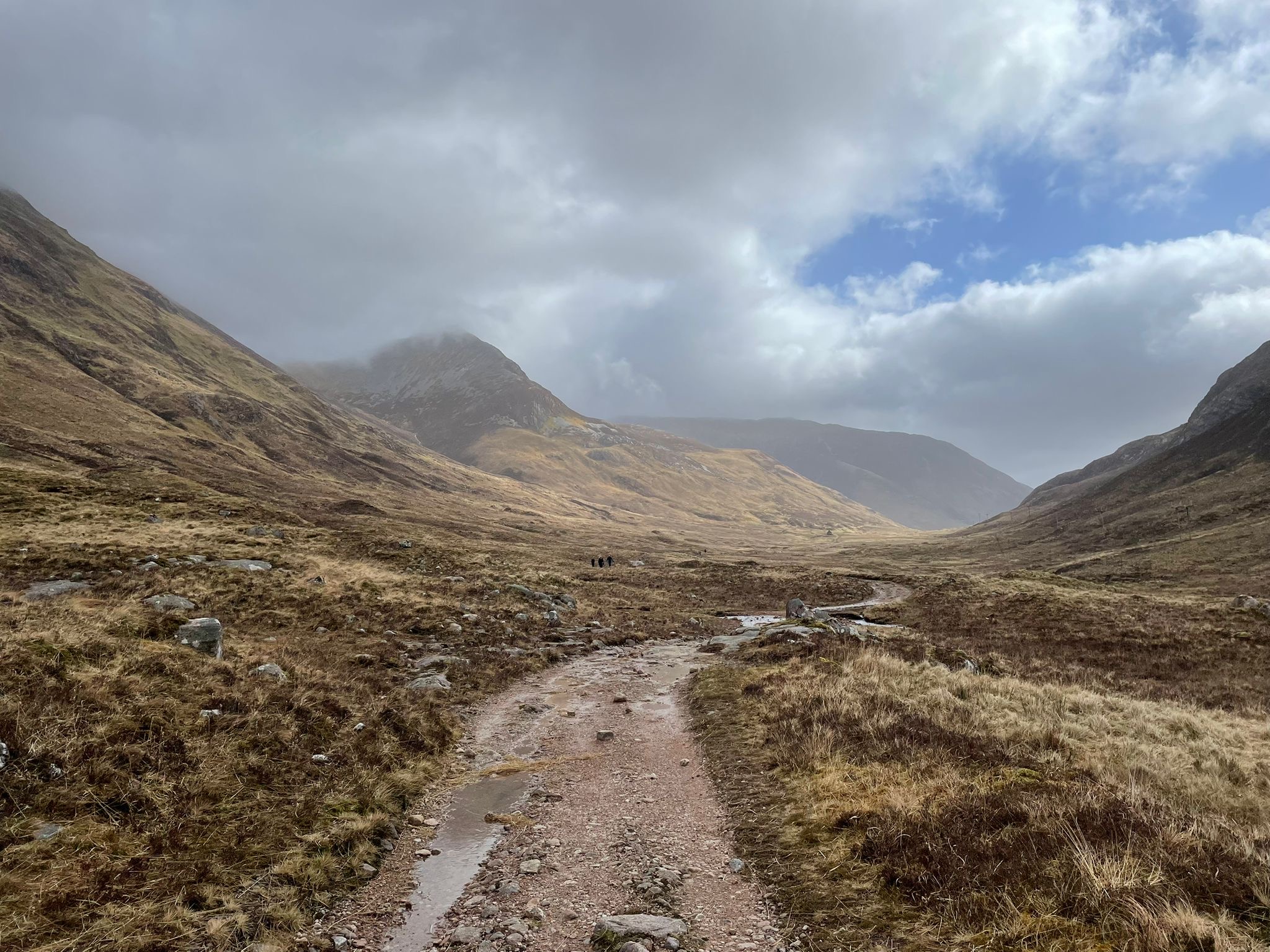 A rocky dirt path winds through a brown grassy valley between rugged hills under a cloudy sky, with patches of blue visible in the distance.