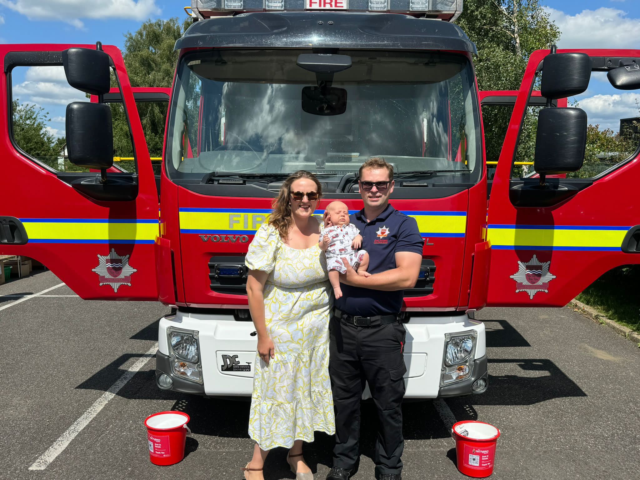 A woman, a man in a fire department uniform holding a baby, stand in front of a fire truck with two red donation buckets on the ground. It’s sunny outside with blue sky and trees in the background.