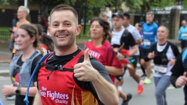 A male runner smiles broadly and gives a thumbs-up during a charity race. He is wearing a red vest with 
