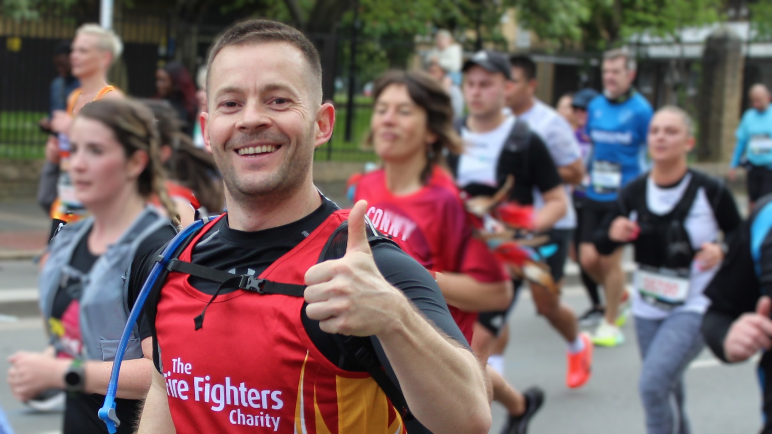 A male runner smiles broadly and gives a thumbs-up during a charity race. He is wearing a red vest with