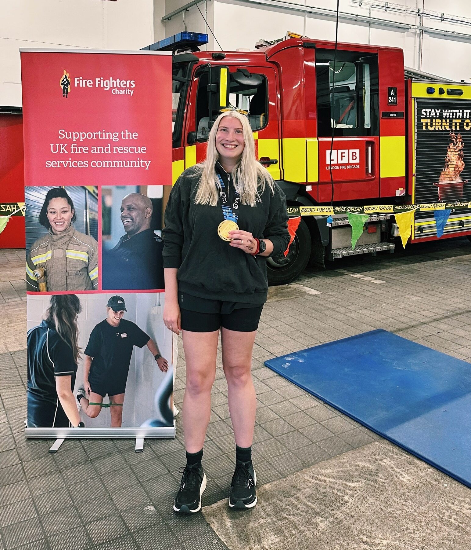 A smiling young woman holds a gold medal and trophy inside a fire station, standing beside a red fire truck and a Fire Fighters Charity banner supporting UK fire and rescue services.
