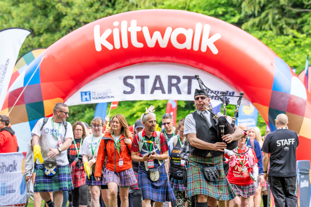 A group of people wearing kilts walk under a large red and white inflatable arch labeled 