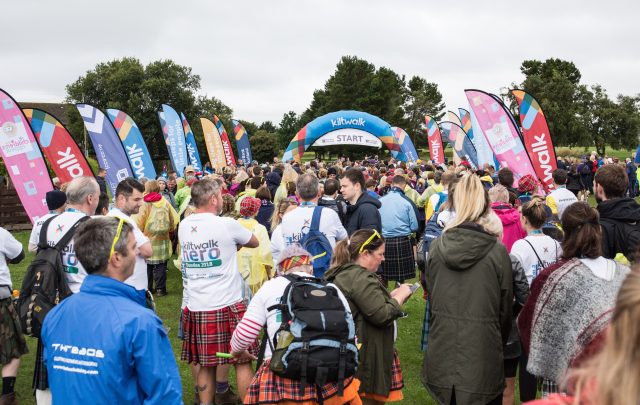 A large crowd of people wearing kilts and event shirts gather at an outdoor starting line under an arch that says 