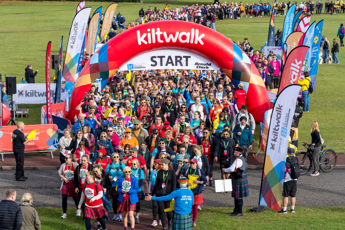 Large crowd of people in colorful outfits, many wearing kilts, gathered at the start line of a Kiltwalk event under a red arch on a grassy field, with banners and flags on both sides.