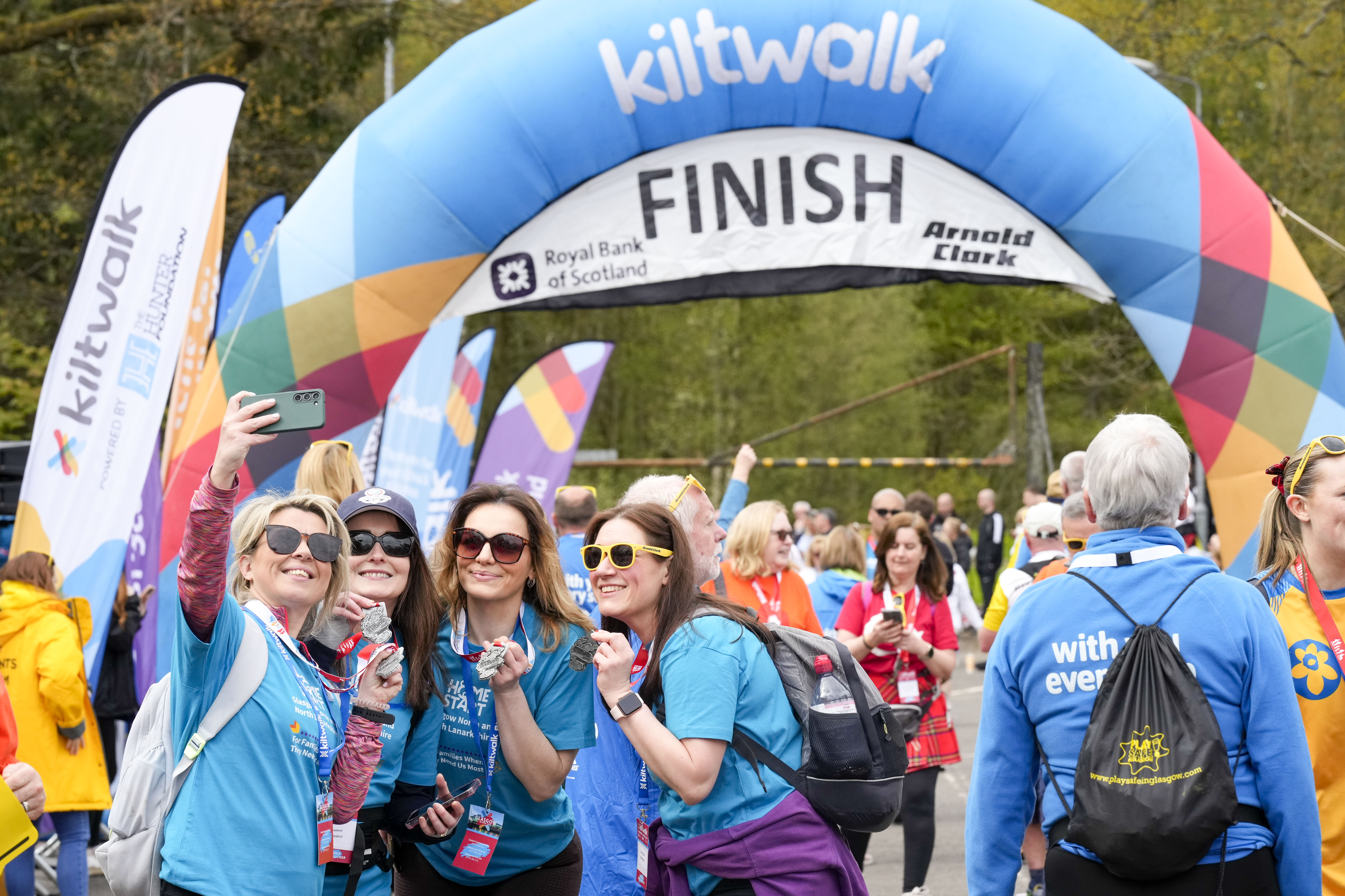 A group of smiling women in blue shirts take a selfie together near the colorful Kiltwalk finish line arch, surrounded by other participants and event banners.