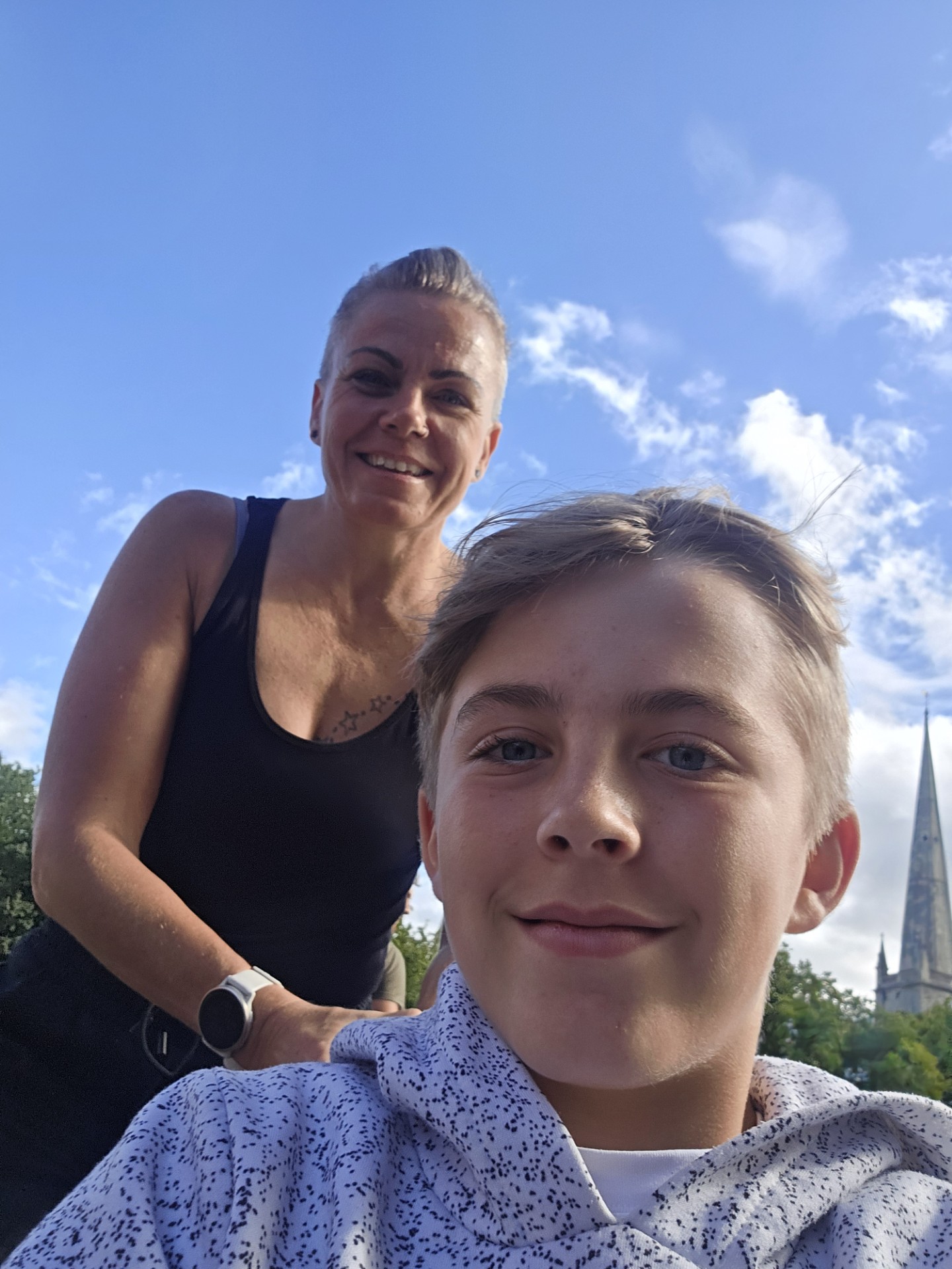 A woman and a boy smiling outdoors under a blue sky with scattered clouds. Trees and part of a tall, pointed church spire are visible in the background.
