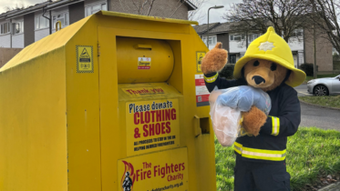 charity mascot blaze bear donating a bag of clothing in the yellow recycling bank outside of a fire station