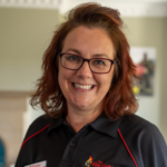 A woman with reddish-brown hair wearing glasses and a black polo shirt with “The Fire Fighters” logo.