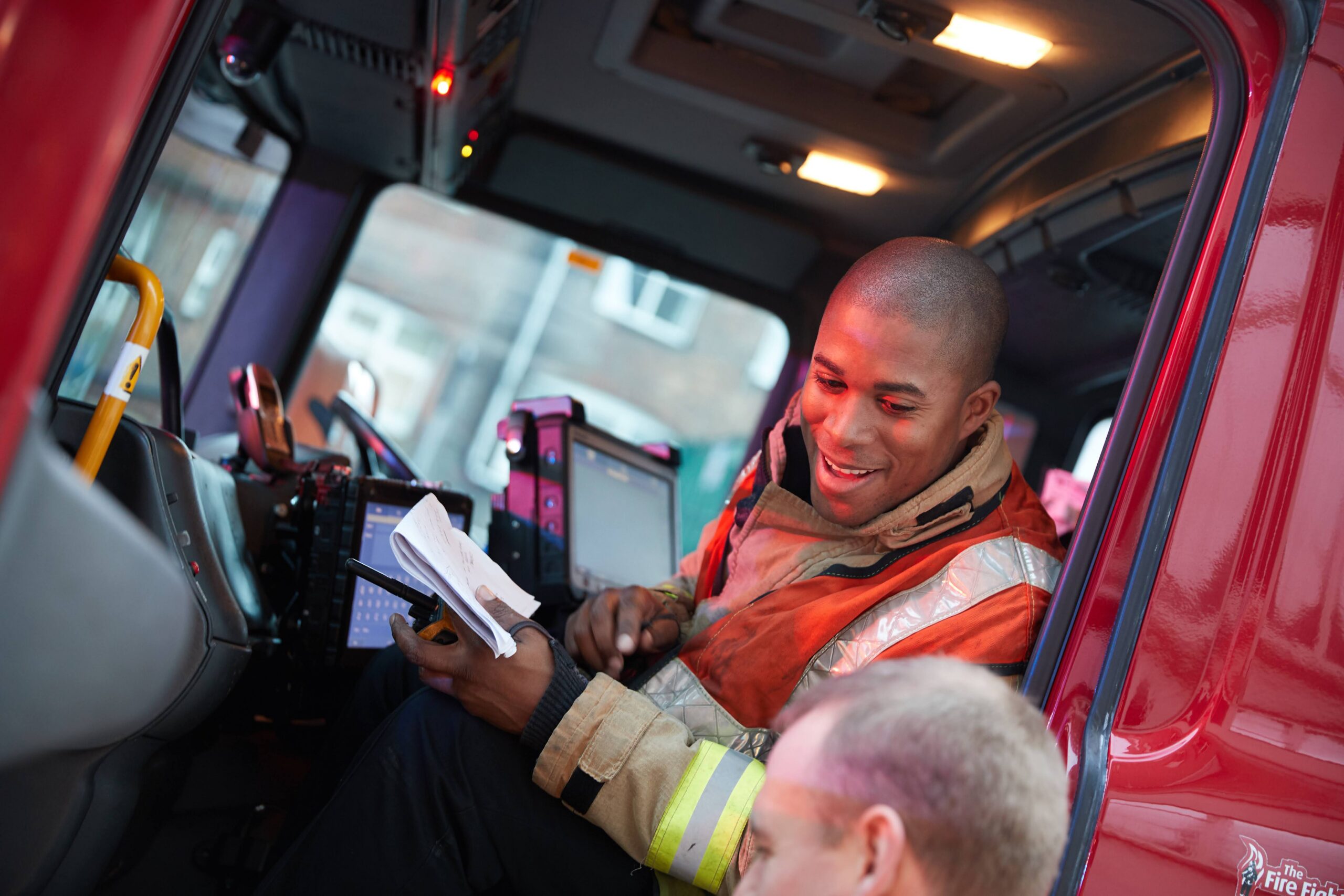 A firefighter in uniform sits inside a fire truck, smiling and holding a notepad, while another person speaks to him from outside the vehicle.