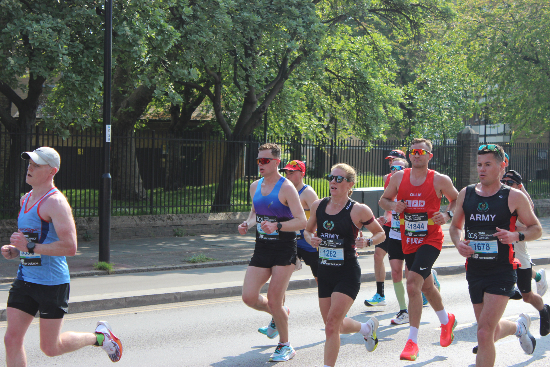 A group of runners participate in a road race on a sunny day, wearing athletic gear and numbered race bibs. Trees and a black iron fence line the street in the background.