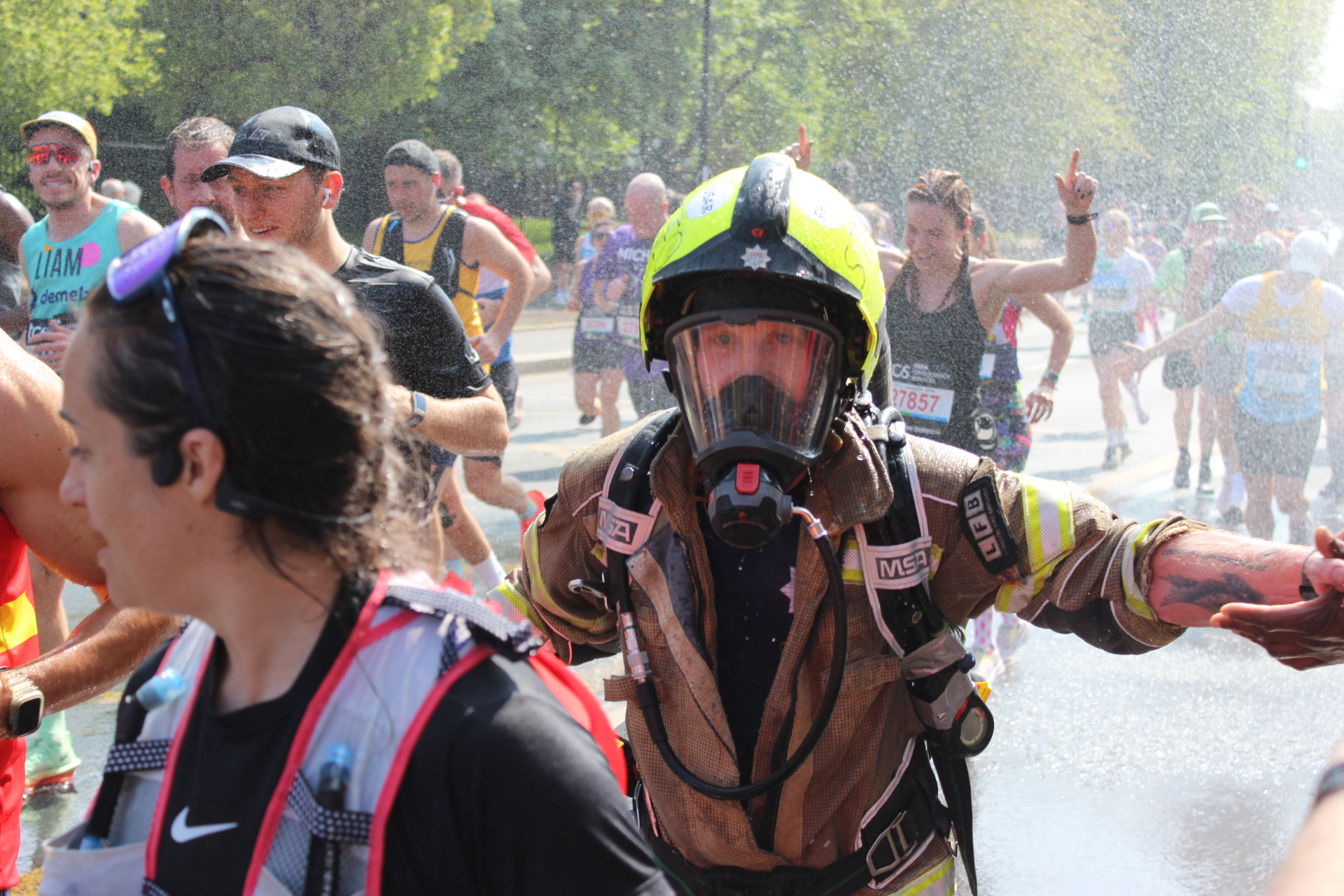 A firefighter in full protective gear and helmet runs among marathon runners, with water spraying in the background on a sunny day. Other runners are visible, wearing race numbers and athletic outfits.