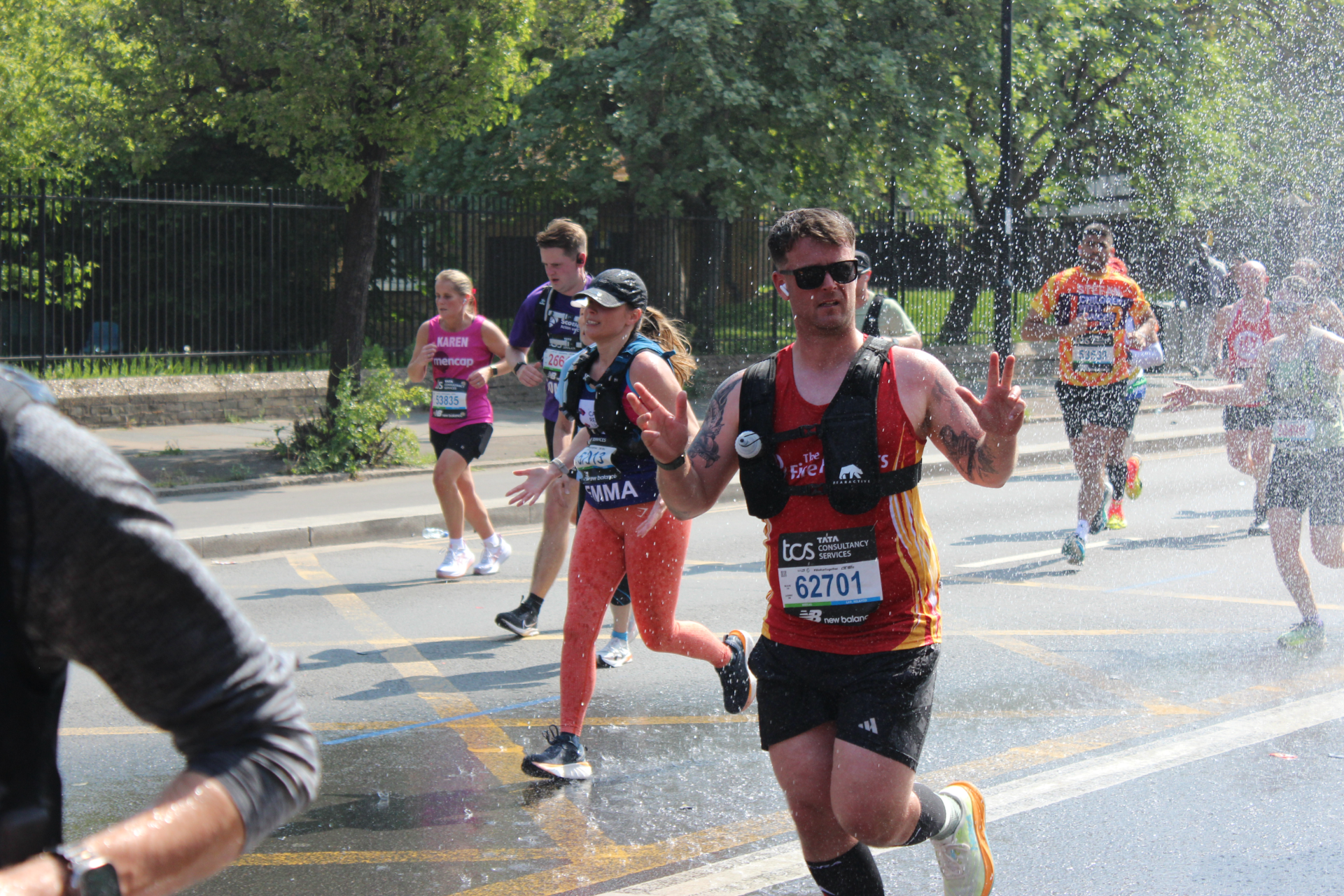A group of marathon runners passes through a spray of water on a sunny day. One runner in sunglasses and a red shirt makes a peace sign with both hands. Other runners are visible in the background.