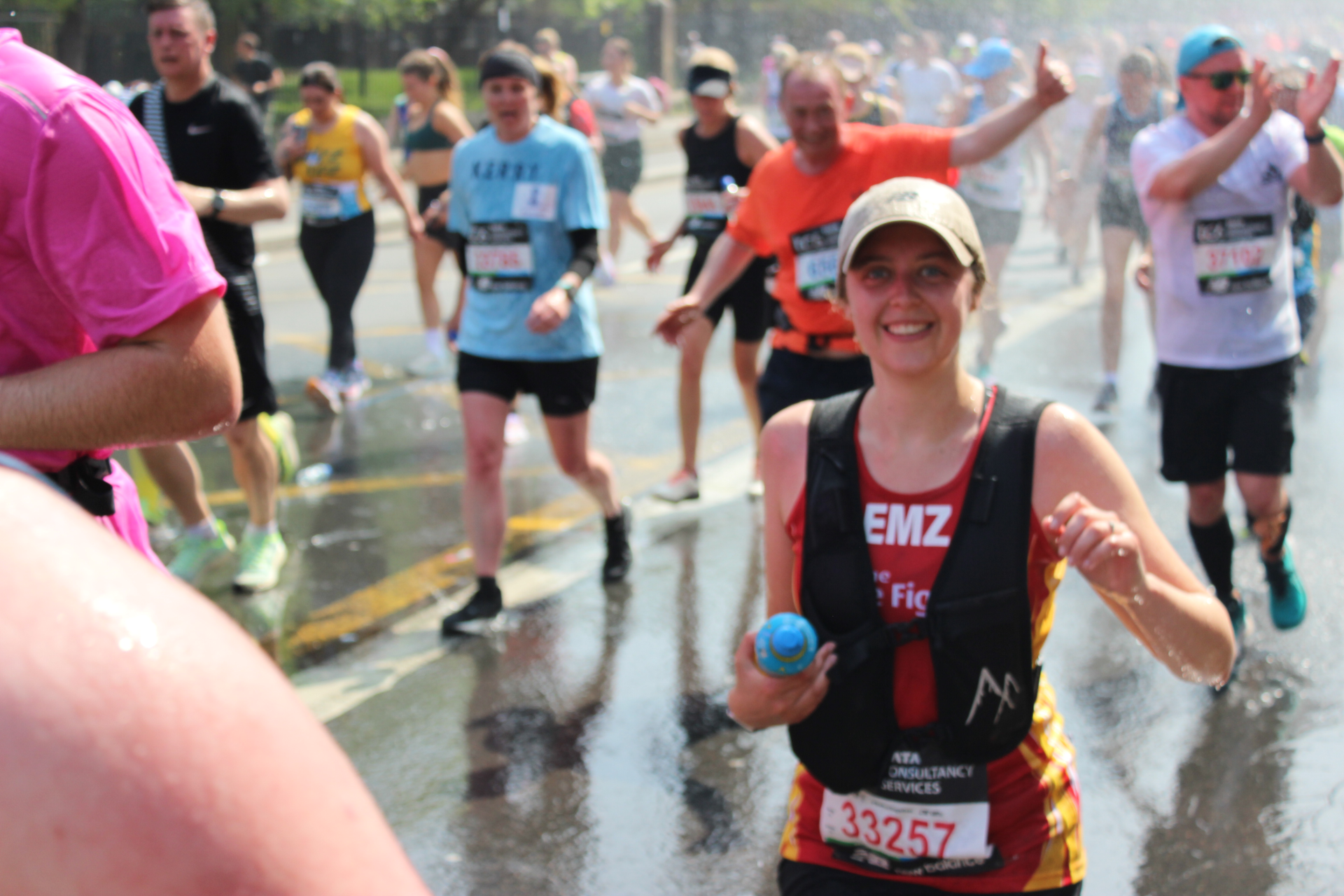 A group of marathon runners, some smiling, run through a mist of water on a sunny day. One woman in the foreground, wearing a tan cap and bib number 3257, smiles and holds a small blue bottle.
