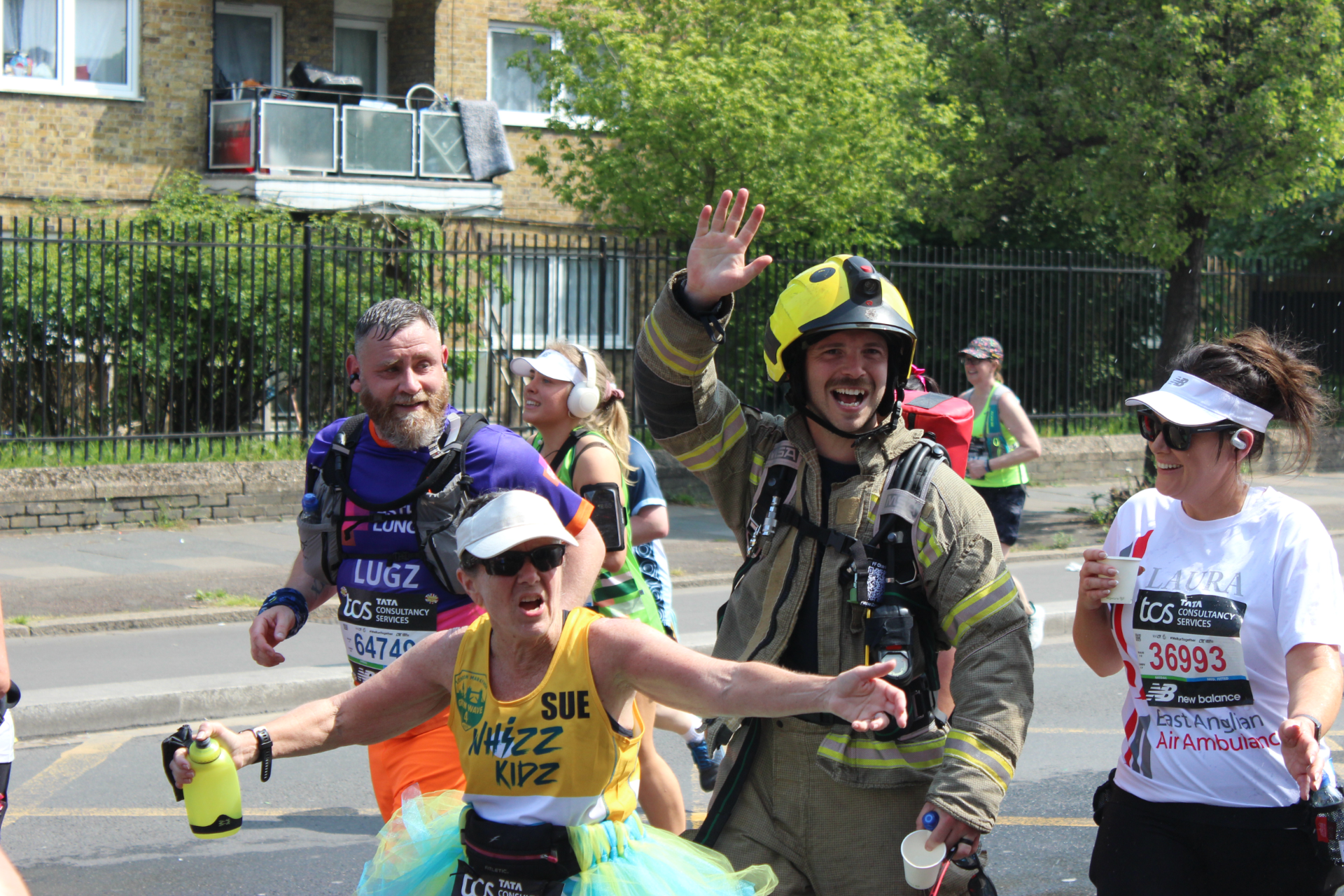 A group of marathon runners, including a smiling firefighter in uniform waving, run together on a sunny day. One woman in front wears a tutu and points, while others appear cheerful and energetic. Houses and greenery are in the background.
