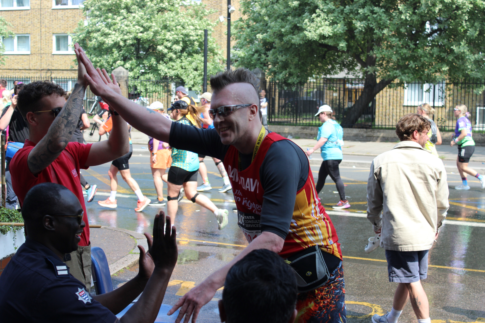 A marathon runner in colorful gear smiles and gives high-fives to supporters at the roadside, while other runners pass by and trees line the street in the background.
