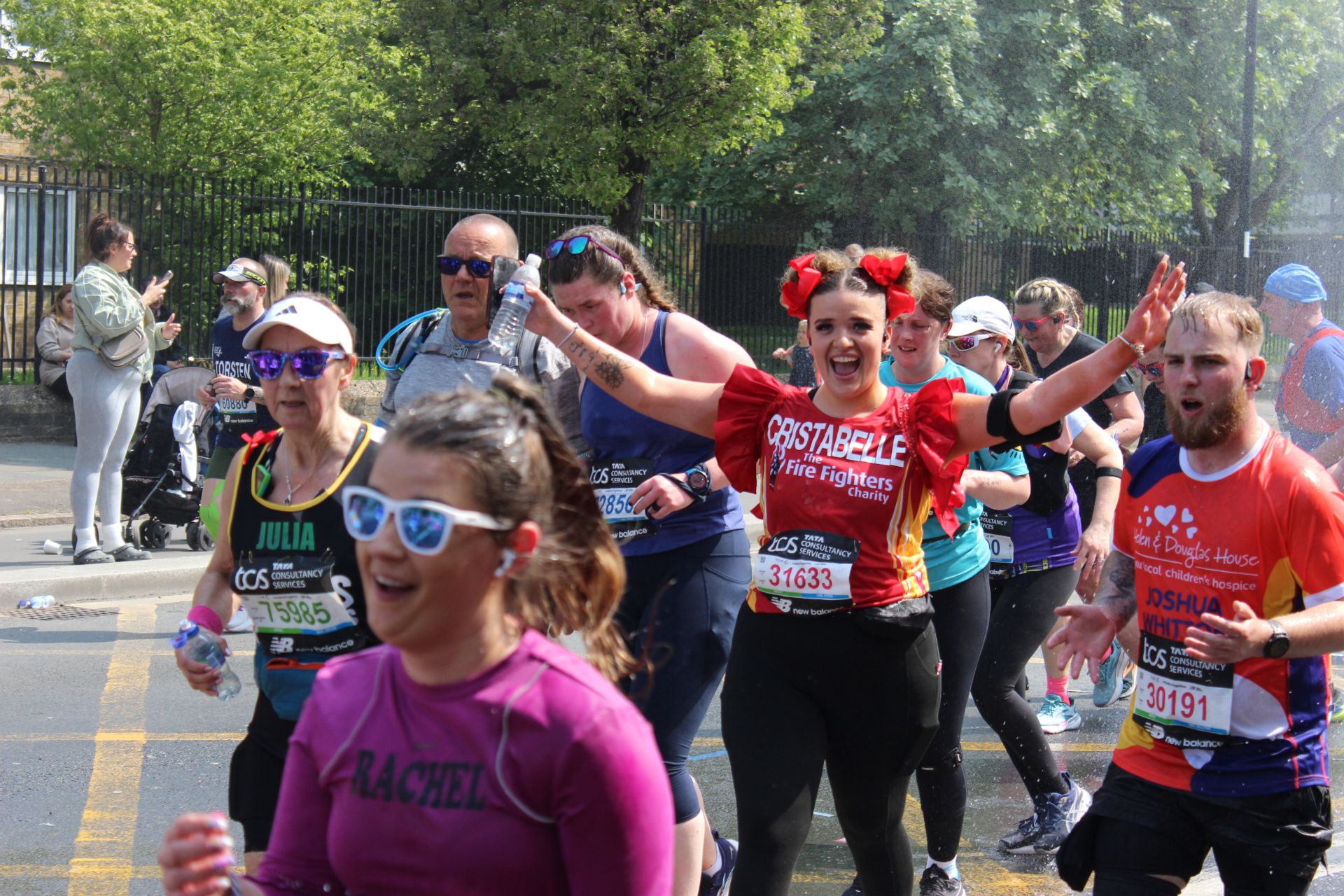 A group of marathon runners in colorful outfits, some smiling and waving at the camera, run down a street on a sunny day. Spectators and trees are in the background.