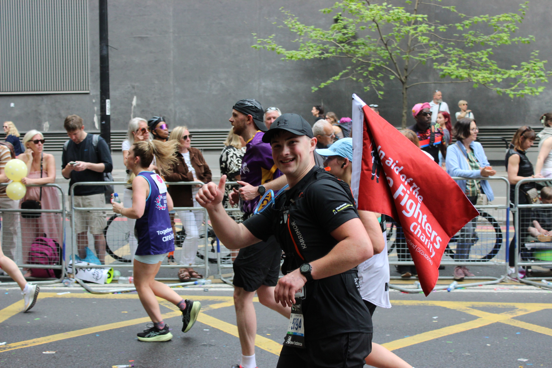 A smiling runner in black athletic gear gives a thumbs up during a marathon. He carries a red flag reading "Fight Cancer," while other runners and spectators line the street behind a metal barrier.