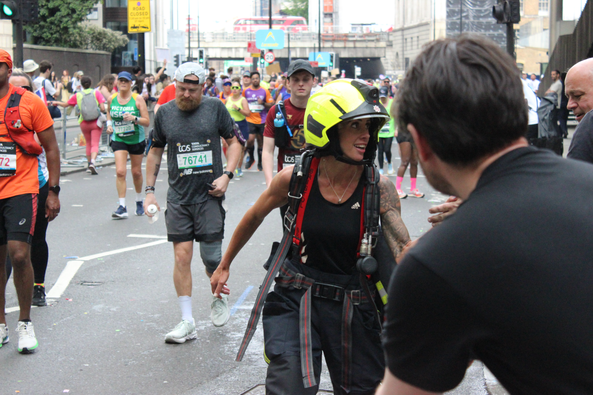 A firefighter in yellow helmet helps marathon runners on a city street, with people running and walking in the background. Some runners wear race bibs and athletic gear. Urban buildings and signs are visible.