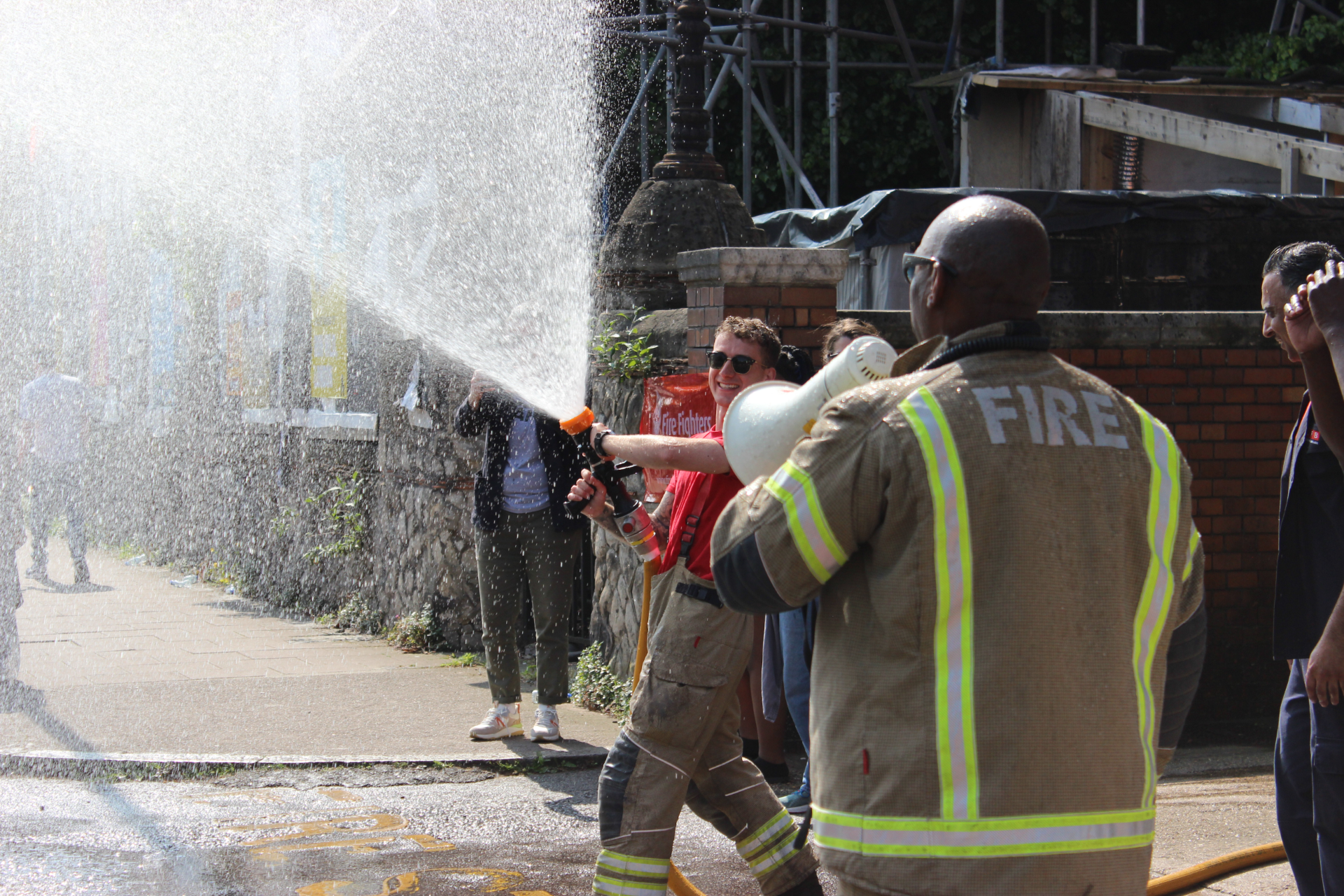 A firefighter helps a person spray water from a fire hose, with water spraying outward. Bystanders watch nearby, and another firefighter stands in the foreground in a jacket labeled "FIRE.