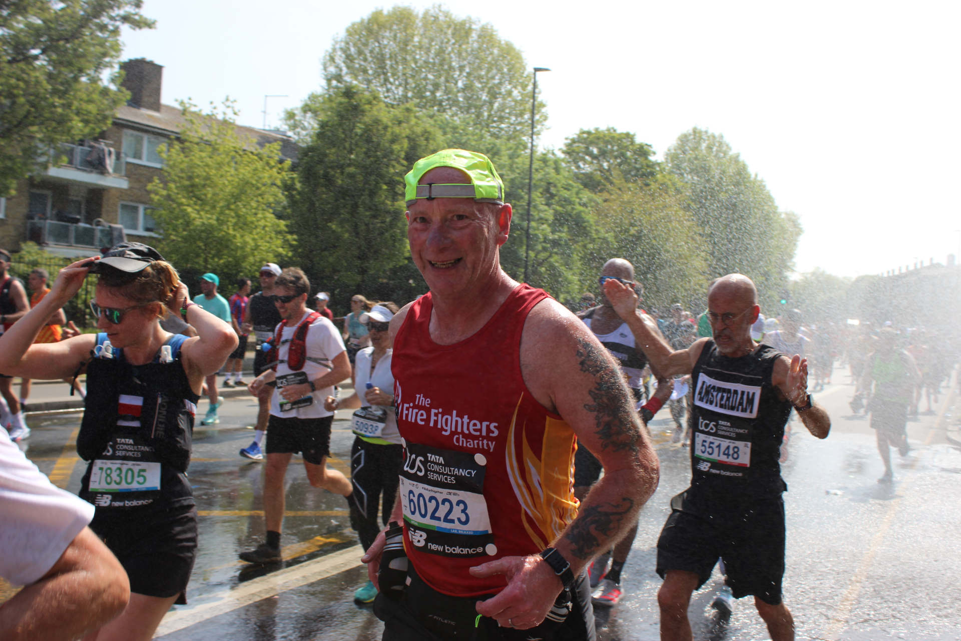 Runners participate in a marathon on a sunny day. A man in a red vest, smiling, is in the foreground, with other runners and a spray of water in the background. Trees and buildings line the street.