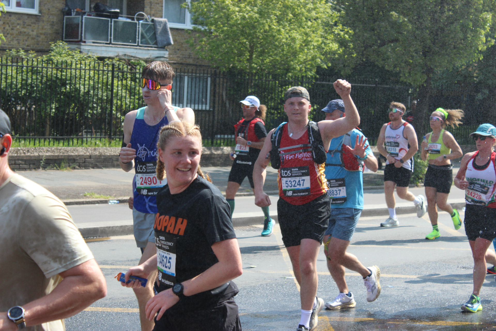 Runners participate in a marathon on a sunny day. Some are smiling and waving while others are focused. Spectators are visible on a balcony in the background. Water droplets are in the air, suggesting a water station nearby.