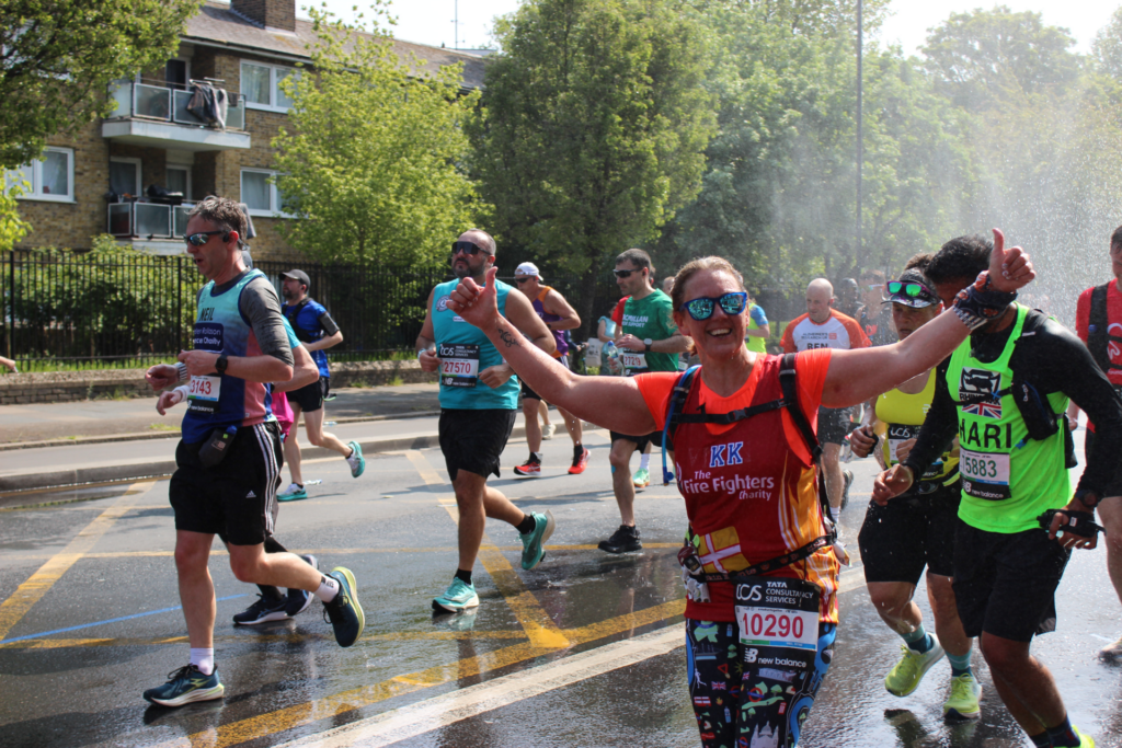 Runners participate in a race on a sunny day. One smiling runner in an orange top gives a thumbs-up toward the camera, while others continue running nearby. Water from sprinklers is visible at the side of the road.