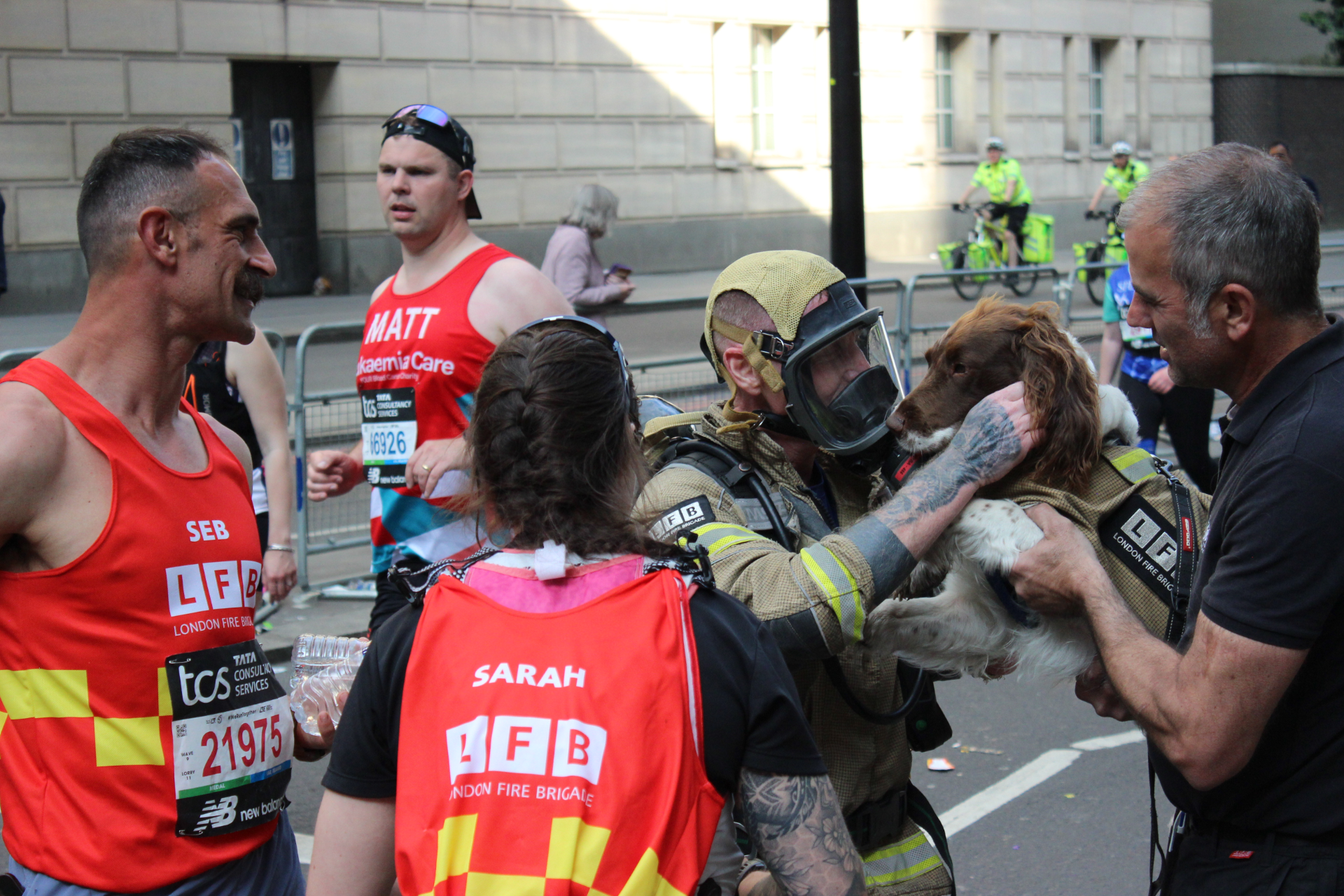 A firefighter in gear gently pets a brown and white dog held by a man, while marathon runners and volunteers in red vests stand nearby. Police officers on bicycles are visible in the background.