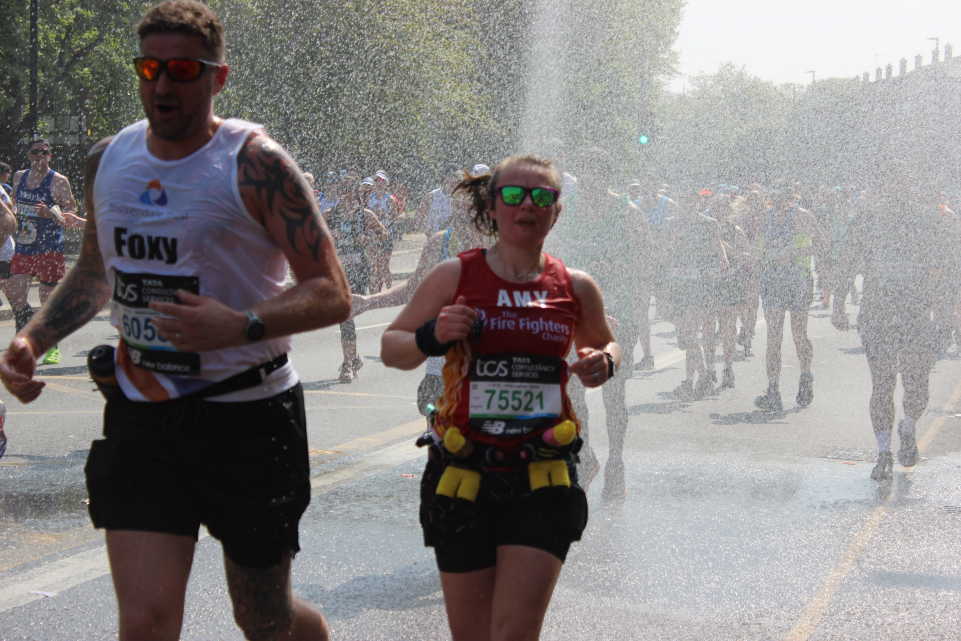 Two marathon runners, one wearing a white shirt labeled "Foxy" and the other in a red shirt, run through a spray of water along a sunny street with other participants in the background.