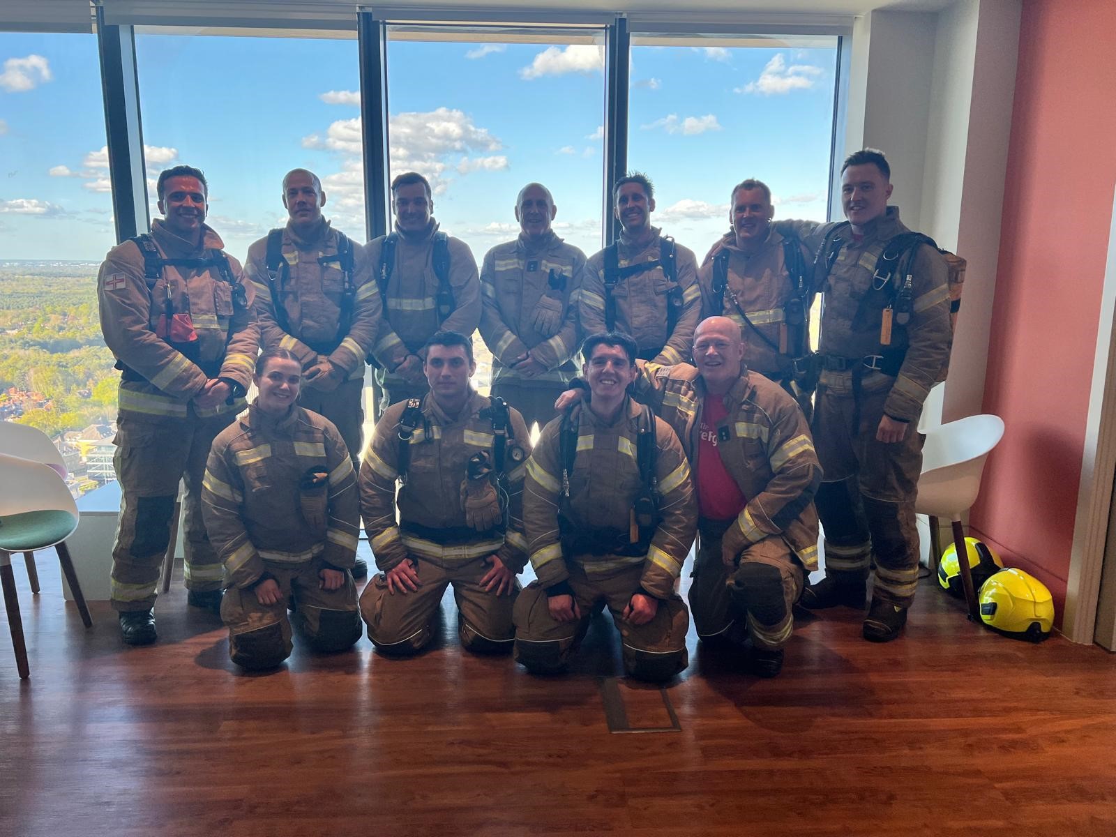 Eleven firefighters in full gear pose together indoors in front of large windows with a scenic view of trees and a blue sky with clouds. Some are standing, others are kneeling, all smiling at the camera.
