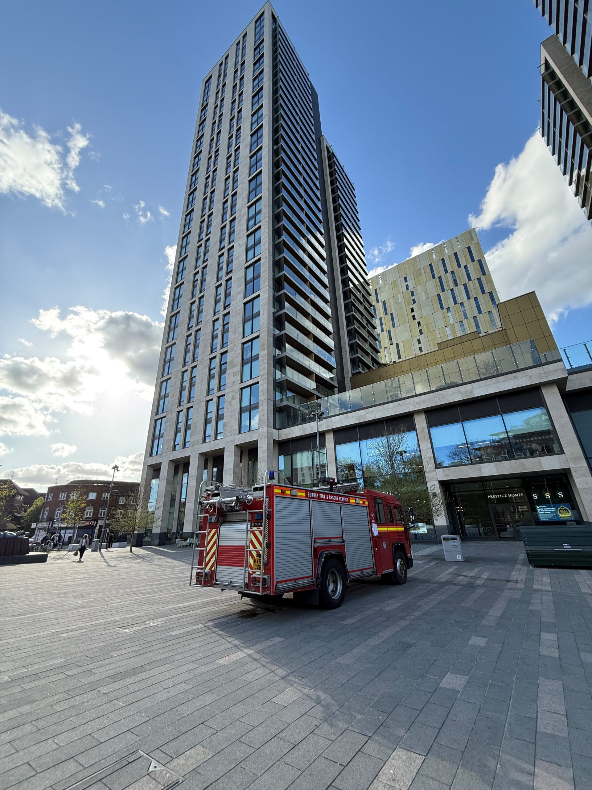 A red fire truck is parked on a wide paved plaza in front of a tall, modern high-rise building with a glass facade, under a blue sky with scattered clouds.