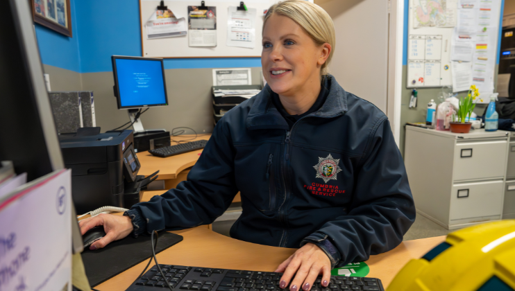 A woman in a Cumbria Fire & Rescue Service jacket sits at a desk, using a computer. She is smiling, with office equipment, documents, and bulletin boards in the background.