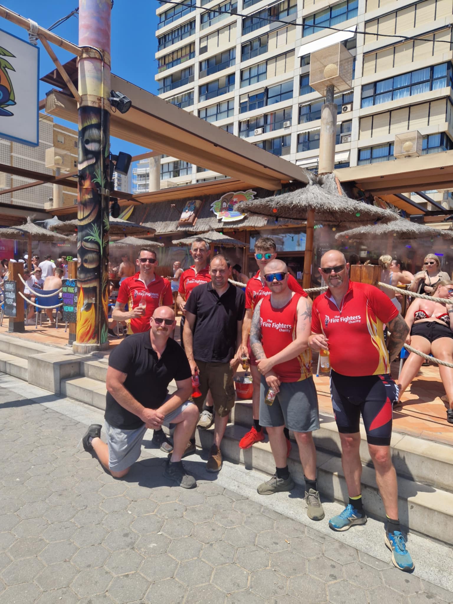 A group of six men, some in matching red cycling jerseys and shorts, pose and smile outside a beachside bar on a sunny day. Tall buildings and people relaxing are visible in the background.