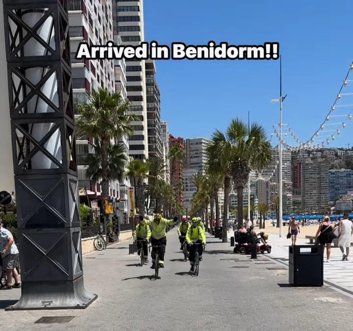 Several police officers on bicycles ride along a sunny promenade lined with palm trees and tall buildings, with people walking nearby and a beach visible to the right. Text reads, "Arrived in Benidorm!!.