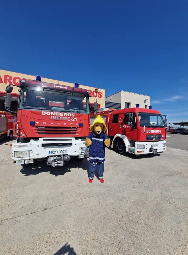 A person in a bear costume wearing a firefighter jacket and helmet stands between two red fire trucks outside a fire station on a sunny day.