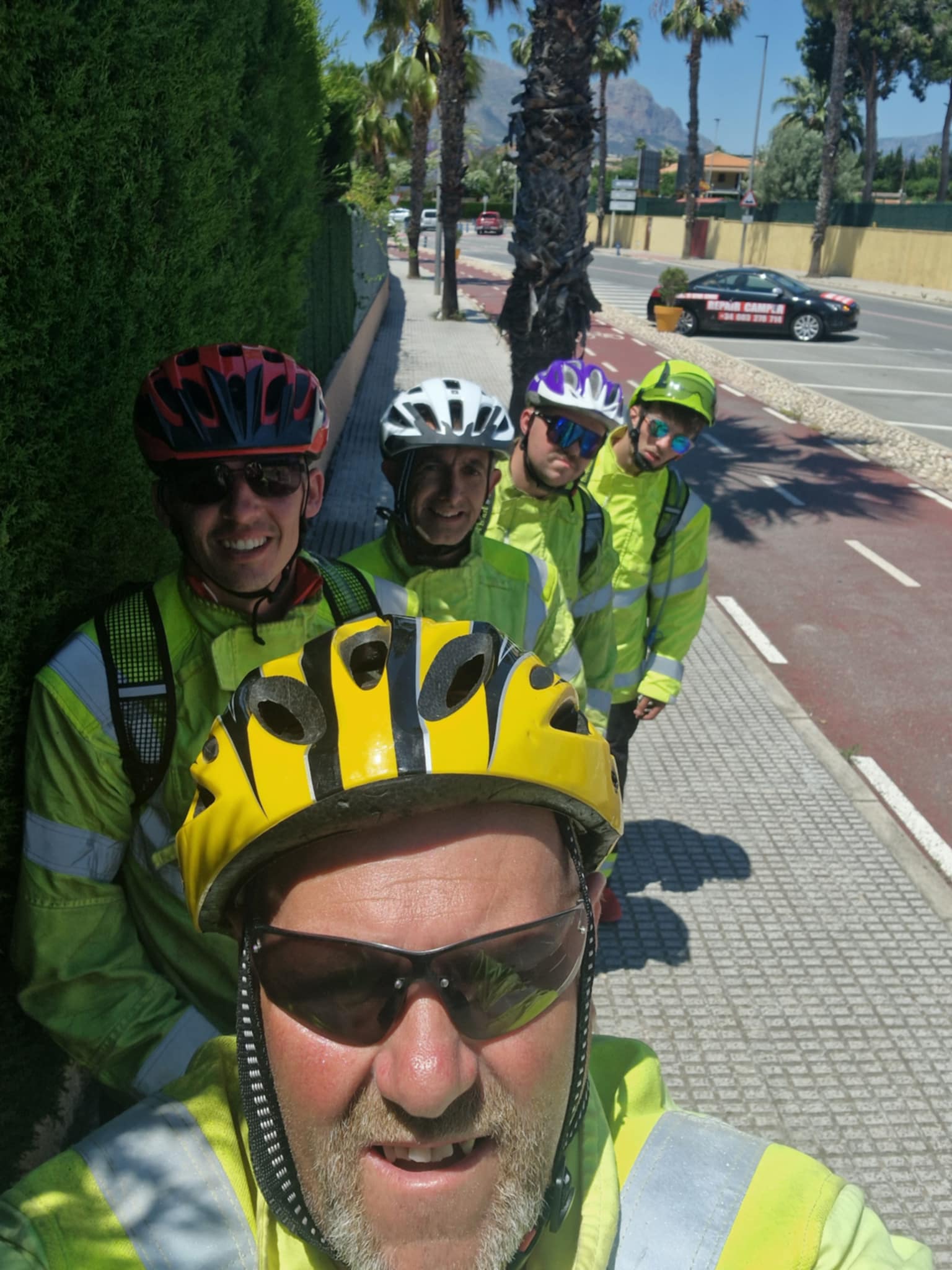 Four cyclists in high-visibility jackets and helmets pose for a selfie on a sunny street lined with palm trees, with mountains visible in the background.