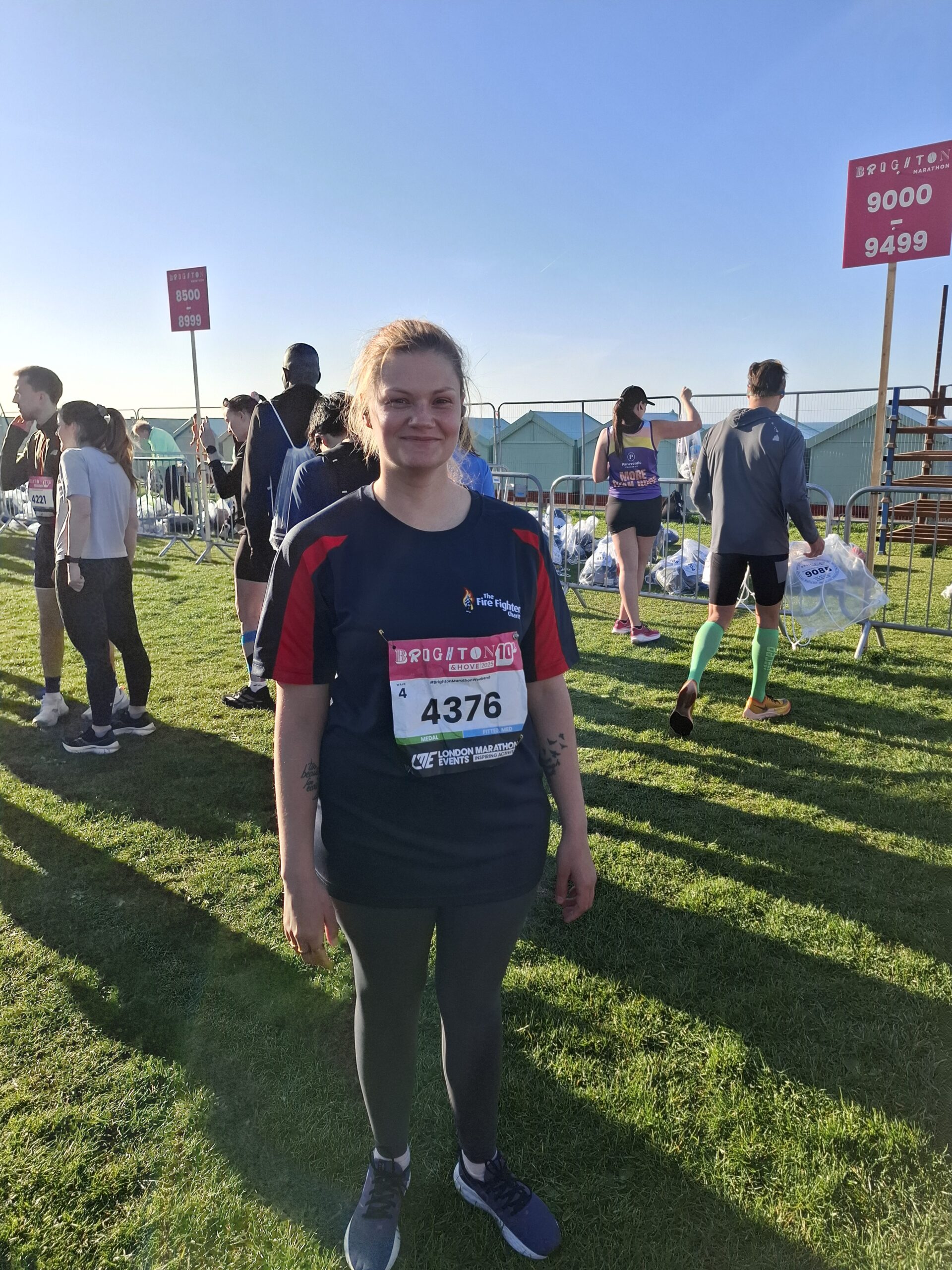 A woman in running gear stands on grass at a race event, smiling at the camera with a race bib numbered 4376. Other runners and signs with start numbers are visible in the background under a clear blue sky.