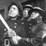 Four vintage firefighters in uniforms and helmets look upward as they work together to hold and aim a fire hose, focused and alert. The image is in black and white.