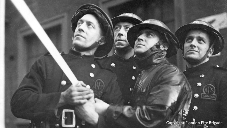 Four firefighters in vintage uniforms and helmets look upward while holding a fire hose. Their expressions are focused and determined. The photo is black and white. Copyright: London Fire Brigade.