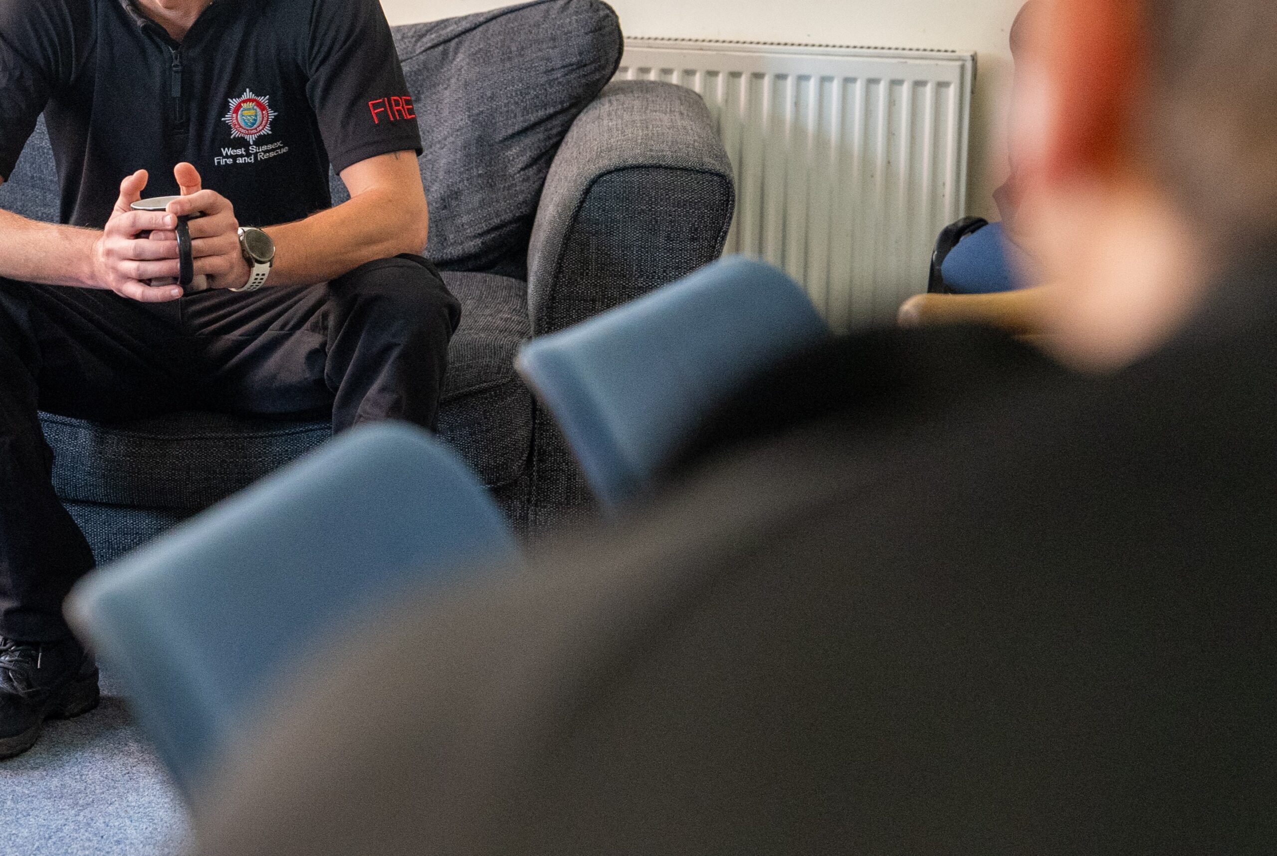 A firefighter in uniform, seated on a couch with hands clasped, is talking to others in a casual indoor setting. Several blue chairs and a radiator are visible; people’s blurred silhouettes are in the foreground.