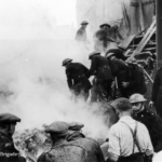 Firefighters and civilians search through smoking rubble after a building collapse in a city street, with debris and damaged structures visible. Several people wear helmets and uniforms. The atmosphere is tense and urgent.