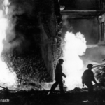 Two firefighters in silhouette walk amid debris and intense flames inside a damaged building at night. The scene is chaotic, with fire illuminating the destruction. Text reads, "Copyright: London Fire Brigade.