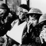 Four exhausted firefighters in uniforms and helmets sit in a row, some with bandaged arms and faces dirty with soot, wiping their eyes and faces, conveying fatigue and distress.