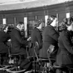 Black and white photo of men and women in uniform sitting at a row of old-fashioned telephone switchboards, wearing headsets and connecting calls. "Copyright London Fire Brigade" appears in the bottom left corner.
