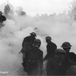 Firefighters and rescue workers in helmets move through thick smoke and debris during an emergency response, with silhouettes of buildings visible in the background. Dense smoke obscures much of the scene.