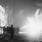 A black and white photo shows firefighters and onlookers silhouetted against a burning building at night, as water is sprayed onto the flames on a debris-strewn street.