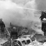 Firefighters in helmets spray water onto heavy smoke and burning rubble, battling a large fire amid collapsed buildings and debris. The scene is chaotic, with dense smoke obscuring parts of the background.
