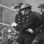 Black and white photo of firefighters in uniform and helmets working together to aim a hose amid smoke and rubble during a fire. Brick walls and debris are visible in the background.