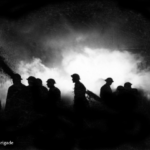Silhouetted firefighters in helmets walk through dense smoke, with bright light illuminating the scene behind them. The mood is tense and dramatic. Copyright: London Fire Brigade.