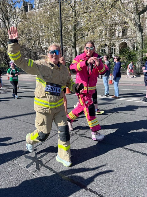 Two smiling women in firefighter suits, one tan and one pink, wave as they run during a race. A race bib is visible, and other runners and spectators are in the background on a sunny day.