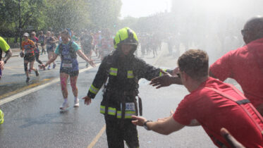 A firefighter in gear runs through mist and high-fives a person in a red shirt during a marathon, with other runners and sprays of water in the background on a sunny day.